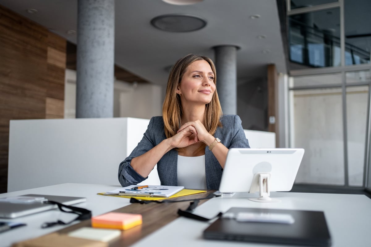 Empresaria sentada en un escritorio moderno, sonriendo y mirando pensativamente hacia el futuro mientras contempla los planes de negocio y las aspiraciones de éxito