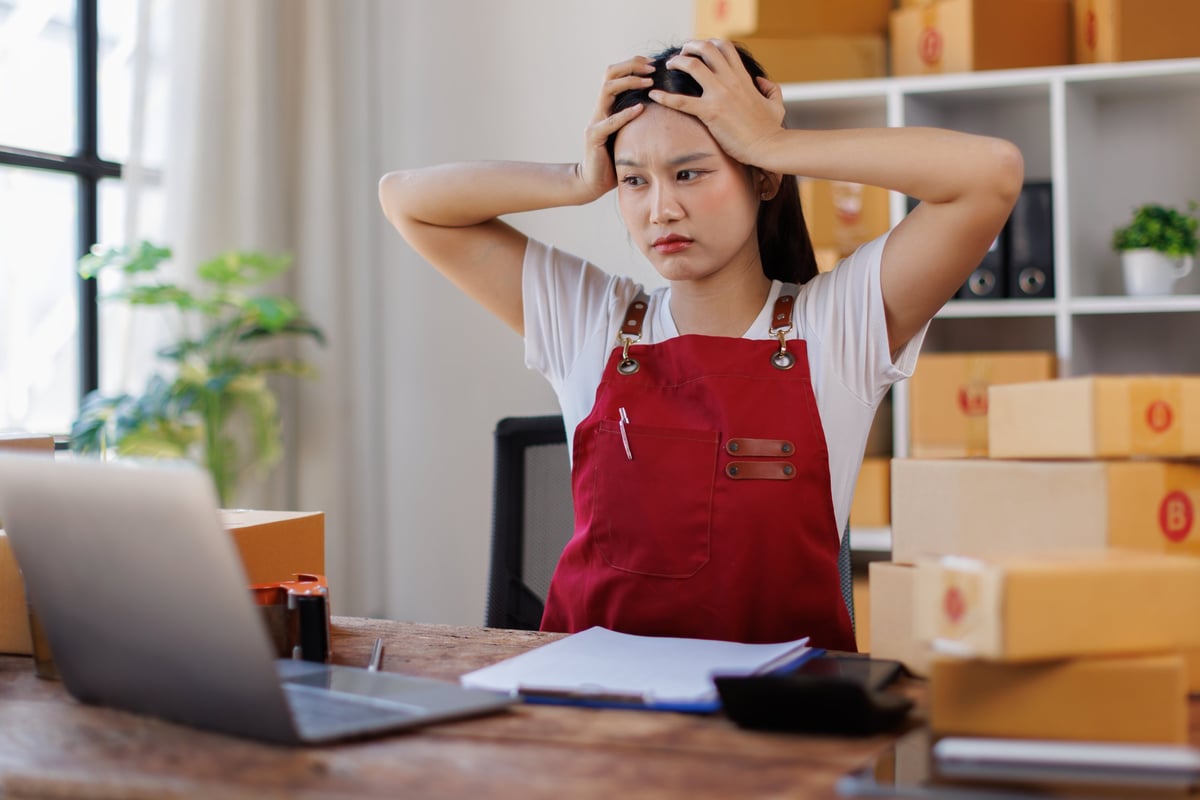Mujer emprendedora sintiéndose abrumada y frustrada, sosteniendo su cabeza entre las manos mientras trabaja en una laptop en un escritorio rodeado de cajas de envío para su negocio de comercio electrónico